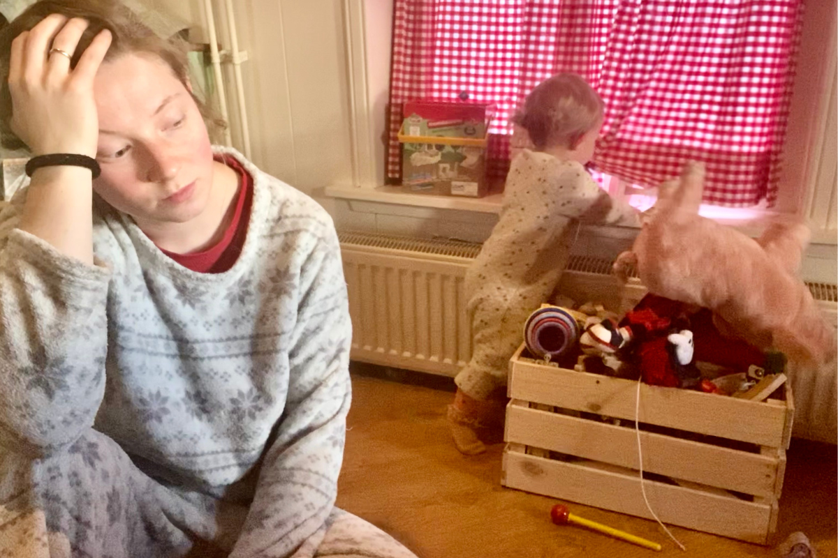 Women in white pjamas sitting on the floor looking depressed with a playing baby in the background